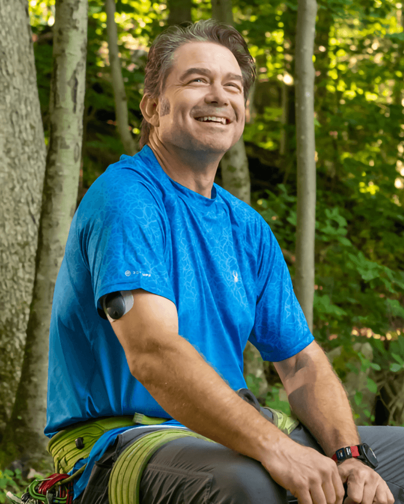 A man wearing a blue shirt and an Eversense glucose monitor smiles while sitting in a sunlit forest.