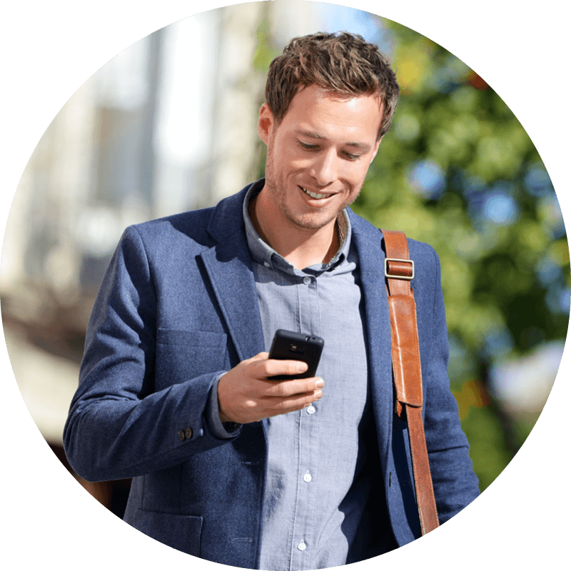 Man smiling, looking at phone, walking outside with blurred greenery and buildings in the background, wearing a blue suit.